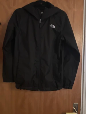 Svart vindjacka från The North Face - Svart vindjacka från The North Face med huva och dragkedja framtill. Jackan har logga på bröstet och ryggen, elastiska muddar och är tillverkad i slitstark syntet med DryVent-teknologi. Perfekt för blåsiga dagar och outdoor-aktiviteter.