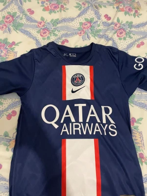 PSG Messi 30 matchtröja Nike blå - Säljer en officiell Paris Saint-Germain t-shirt med Messi 30 på ryggen. Tröjan är marinblå med vita och röda detaljer, PSG-logga och Qatar Airways tryck på framsidan. Tillverkad i polyester med korta ärmar, perfekt för fotboll och sport.