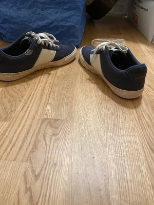 Mörkblå och vita sneakers med snörning - Snygga sneakers i mörkblått och vitt med klassisk snörning och rund tå. Skorna har en vit sula och detaljer med röd söm på hälen. Materialet är textil och syntet, vilket gör dem lätta och bekväma. Perfekt för dig som gillar stilrena och sportiga skor.