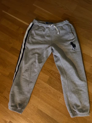 Polo Ralph Lauren mjukisbyxor pris ej hugget - Snygga grå mjukisbyxor från Polo Ralph Lauren med klassisk marinblå logga broderad på benet. Byxorna har vita och marinblå ränder längs sidorna, snörning i midjan och mjukt bomullsmaterial. Endast använda 1 gång har växt ur dom. Perfekt för om man vill vara bekväm men ändå hålla stilen🙌