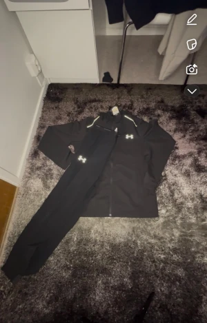 Svart träningsset från Under Armour - Snyggt svart träningsset från Under Armour med jacka och byxor. Jackan har dragkedja framtill och reflekterande detaljer vid axlarna. Både jacka och byxor har Under Armour-loggan i vitt. Perfekt för gymmet eller löprundan.