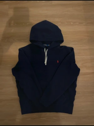 Marinblå hoodie från Polo Ralph Lauren - Snygg marinblå hoodie från Polo Ralph Lauren med klassisk huva och vita dragsnören. Broderad röd logga på bröstet. Mjuk bomullskänsla och avslappnad passform, perfekt för en chill look.