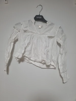 Vit blus med volang från Gina Tricot - Söt vit blus från Gina Tricot i storlek 134/140. Blusen har långa ärmar med smock vid ärmslut, volangdetaljer vid axlarna och spetsinslag över bröst och rygg. Tillverkad i ett lätt och luftigt tyg, perfekt för en trendig look.