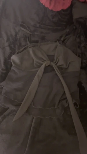 Svart croppad linnetopp med knyt - Säljer en svart croppad linnetopp med smala axelband och snygg knytning framtill. Toppen har vågiga kanter och är tillverkad i ett lätt, syntetiskt material som känns skönt mot huden. Perfekt för dig som gillar trendiga och stilrena plagg.