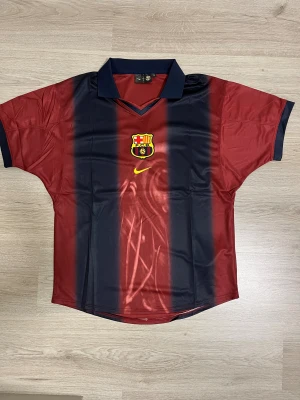 Barça x Cactus Jack Nike t-shirt - Säljer en limited edition FC Barcelona t-shirt i samarbete med Cactus Jack och Nike. Tröjan har klassiska röda och mörkblå ränder, v-ringad krage och tryck med Barça-logga fram samt Cactus Jack, nummer 2 och BARÇA bak. Materialet är glansig polyester, perfekt för fotbollsfans.