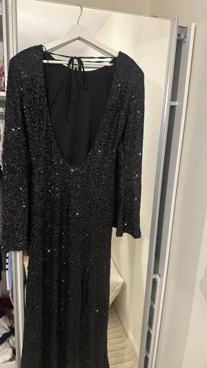 Svart glittrig långklänning L - Svart långklänning täckt av glittrande paljetter med djup urringning i ryggen och knytband i nacken. Klänningen har långa ärmar och en rak, elegant siluett. Perfekt för dig som vill sticka ut med en maxiklänning som verkligen glittrar.