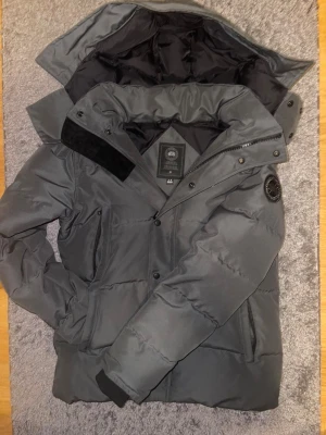 Mörkgrå dunjacka från Canada Goose - Snygg mörkgrå dunjacka från Canada Goose med huva, svarta detaljer och patch på ärmen. Jackan har dragkedja och knappar framtill, samt två sidofickor. Perfekt för kalla dagar och har en clean, modern look.