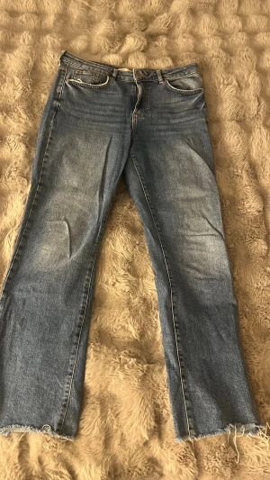 Blå raka jeans från Gina Tricot strl 40 - Säljer ett par klassiska blå jeans från Gina Tricot, The Good Project. Jeansen har raka ben, normal midja och råa, fransiga benslut. Tillverkade i mer hållbar bomull. Perfekta för en avslappnad och trendig look.