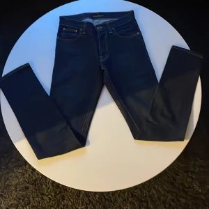 Mörkblå Nudie Jeans 30/34  - Snygga mörkblå jeans från Nudie Jeans i modellen slim fit med klassisk femficksdesign och orange kontrastsömmar. Ej använda mer är 3ggr. Jeansen har raka ben och coola broderade detaljer på bakfickorna. Perfekt för dig som gillar stilrena och moderna jeans.