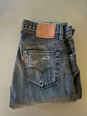 Levi's 501 mörk grå jeans W29 L32 - Mörkgråa Levi's 501 jeans med klassisk femficksdesign och slitna detaljer på bakfickan. Jeansen har rak passform och är tillverkade i robust denim med tydliga kontrastsömmar. Perfekt för dig som gillar en tidlös och cool look.