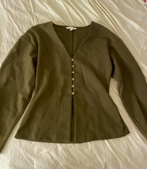 Olivgrön blus med v-ringning och guldknappar - Snygg olivgrön blus med v-ringad hals och guldfärgade knappar framtill. Blusen har långa ärmar och en figurnära passform. Endast testad.