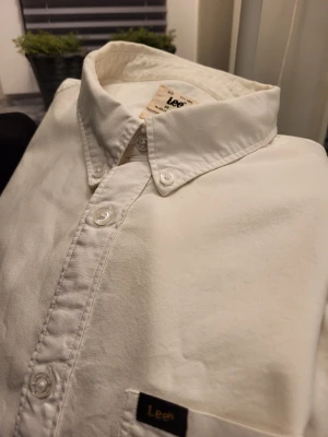 Vit skjorta från Lee i bomull - Klassisk vit skjorta från Lee i regular fit med button-down krage och bröstficka. Skjortan är tillverkad i bomull och har långa ärmar samt knappar framtill. Perfekt för en clean och stilren look.