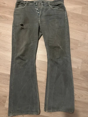Grå bootcut jeans från Acne Studios - Säljer ett par gråa jeans från Acne Studios med bootcut passform och knappar i gylfen. Jeansen har slitningar och hål på framsidan samt klassiska bakfickor. Materialet är 100% bomull och de har en cool, lite vintage vibe. Pris kan diskuteras 