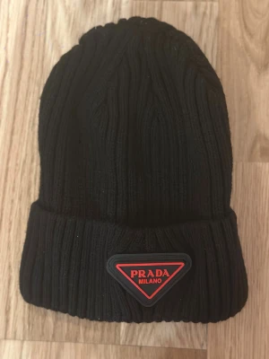 Svart ribbad mössa  - Svart ribbstickad mössa med uppvikt kant och ikonisk röd logotyp framtill. 