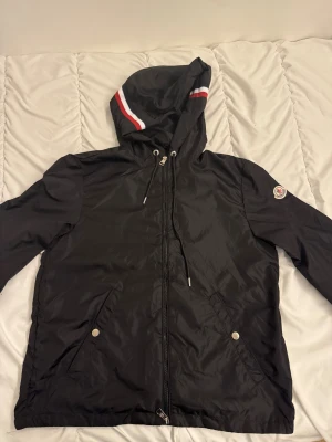 Svart vindjacka från Moncler - Svart vindjacka från Moncler med huva som har vita och röda ränder. Jackan är tillverkad i 100% polyester och har dragkedja framtill, snörning vid huvan och två fickor med tryckknappar. Moncler-logga på vänster ärm och insida.