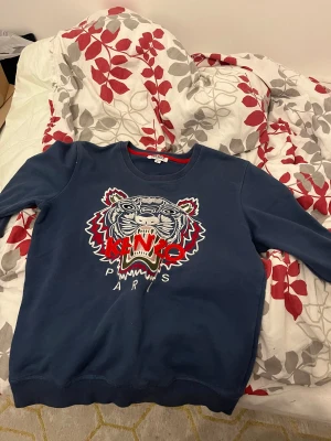 Mörkblå Kenzo sweatshirt med tiger - Säljer en mörkblå sweatshirt från Kenzo i storlek L med ikonisk broderad tiger och röd Kenzo-logga på bröstet. Tröjan har rund hals, långa ärmar och ribbade muddar. Perfekt för dig som vill sticka ut med ett coolt statement-plagg.