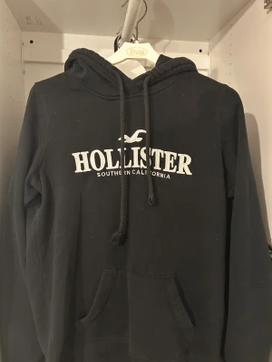 Svart hoodie från Hollister - Svart hoodie från Hollister med vit broderad logga och text på bröstet. Tröjan har huva med snörning, känguruficka och långa ärmar. Perfekt för chill dagar och har en klassisk streetstil. Materialet är mjuk bomull och polyesterblandning. Hör av er vid minsta fundering!