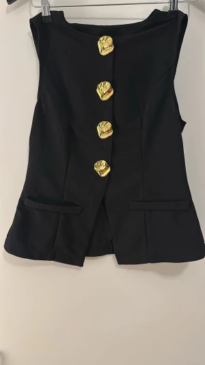 Svart kavajväst från SHEIN med guldknappar - Svart kavajväst från SHEIN i storlek S, med unika, skulpturala guldknappar framtill. Västen har en stilren, figurnära passform och två dekorativa fickor. Perfekt för dig som vill sticka ut med coola detaljer och modern look.