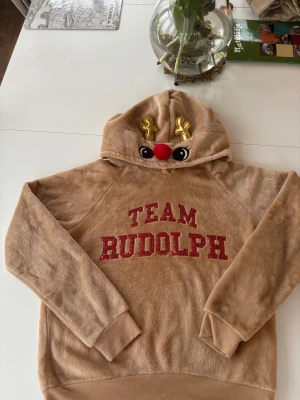 Brun Team Rudolph hoodie med huva - Mjuk och fluffig brun hoodie med röd text 'TEAM RUDOLPH' framtill. Huvan har roliga detaljer som ögon, en röd nos och gyllene horn, vilket ger ett renmotiv. Långa ärmar och ribbade muddar. Perfekt för dig som gillar julmys.
