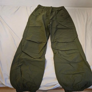 Gröna cargopants - Säljer ett par gröna cargopants med breda ben och resår både i midjan och vid bensluten. Byxorna har stora fickor med lock och knapp framtill, vilket ger en cool och avslappnad streetstil. Har ett polyester aktigt tyg