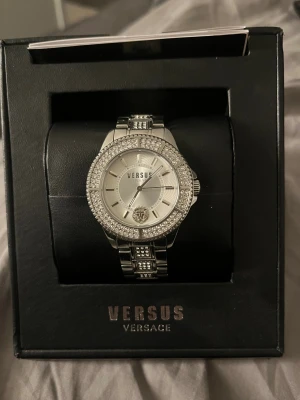 Versus Versace silverfärgad klocka - Snygg klocka från Versus Versace i silverfärgat rostfritt stål med glittrande stenar runt urtavlan och på länken. Rund urtavla med minimalistiska markeringar och Versus-logga, samt ett lejonhuvud vid 6-tiden. Länkarmband med dekorativa detaljer.