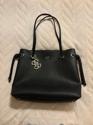 Svart handväska från Guess - säljer en väska från Guess med guld detaljer. Den har ett fack på baksidan med drag kedja och ett fack på innersidan av väskan. Den är i väldigt bra skick och är i skinn. måtten är ungefär 40x30x15