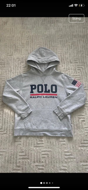 Grå hoodie Polo Ralph Lauren M - Grå hoodie från Polo Ralph Lauren med stor logga på bröstet och amerikansk flagga på ena ärmen. Mjuk insida i fleece, känguruficka och huva. Snygg och sportig stil, perfekt för chill dagar. Materialet är bomullsblandning. storlek M barn 
