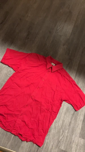 Röd kortärmad skjorta från Clique XL - Riktigt snygg röd skjorta från Clique i storlek XL. Skjortan har korta ärmar, klassisk krage, bröstficka och knäppning framtill med matchande röda knappar. Perfekt för dig som vill sticka ut med färg. Materialet är mjukt och känns skönt mot huden.