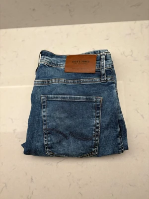 Blå jeans från Jack & Jones - Snygga blå jeans från Jack & Jones med klassisk femficksdesign och brunt läderpatch bak i midjan. Jeansen har kontrastsömmar och är tillverkade i slitstarkt denimtyg. Perfekt för dig som gillar en tidlös och avslappnad stil.