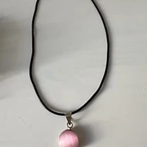 Halsband med rosa pärlhänge - Svart halsband med ett runt, glansigt rosa pärlhänge. Enkelt och stilrent smycke som passar perfekt för att ge din outfit en söt touch. Hängsmycket har en metallögla och halsbandet är i tunn, svart snodd.