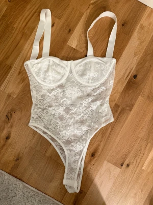 Vit spetskorsett body - Säljer en vit korsett body med breda axelband och vacker spets över hela plagget. Den har bygelkupor för extra stöd och en feminin, figurnära passform. Perfekt för att skapa en snygg och trendig look.