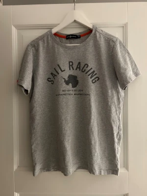 Grå t-shirt Sail Racing M - Grå t-shirt från Sail Racing med tryck på bröstet som visar 'Sail Racing Antarctica Expedition' och en karta över Antarktis. Klassisk rund hals och korta ärmar. Skön och mjuk i bomull, perfekt för en avslappnad stil.