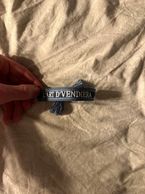 Blå tygarmband L'ART D'VENDIERA - Snyggt blått tygarmband med vit broderad text 'L'ART D'VENDIERA'. Armbandet har flätade band och tofsar i ändarna för att knyta runt handleden. Perfekt för dig som gillar unika accessoarer med cool design.