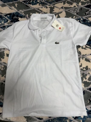 Vit polotopp från Lacoste XS - Säljer en klassisk vit polotopp från Lacoste i storlek XS. Toppen har krage, korta ärmar och knappar framtill. Materialet är bomull och den har den ikoniska gröna krokodilloggan broderad på bröstet. Perfekt för en clean och sportig look.