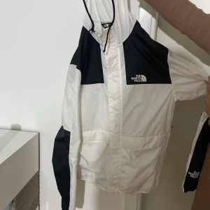 Svartvit vindjacka från The North Face - Säljer en snygg svartvit vindjacka från The North Face med huva och dragkedja framtill. Jackan har logga på bröstet och ärmarna, samt praktiska fickor. Perfekt för dig som gillar streetstyle och vill ha en lätt jacka med cool design.