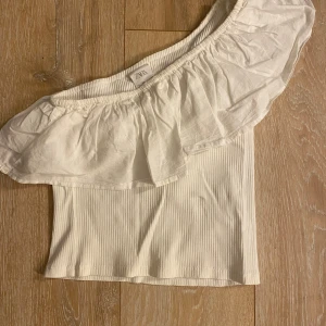 Vit oneshoulder topp från Zara - Säljer en vit ribbad topp från Zara med oneshoulder-design och bred volang längs halsringningen. Toppen är ärmlös och har en snygg, croppad passform som passar perfekt till jeans eller kjol.