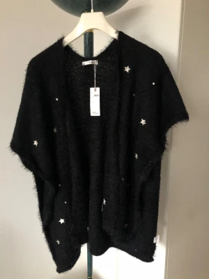 Svart stickad kofta med stjärnor - Svart, fluffig och stickad kofta från Mimi med korta ärmar och silverfärgade stjärnor som detaljer. Koftan har en loose passform och är perfekt att slänga över en t-shirt för en chill look. Materialet är mjukt och ger en mysig känsla.