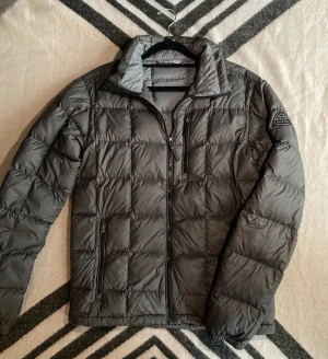 Pufferjacka från McKinley - Snygg grå pufferjacka från McKinley med quiltad design och hög krage. Jackan har dragkedjor framtill och på fickorna, samt en logga på ärmen. Perfekt för kalla dagar och har en modern, boxig look.