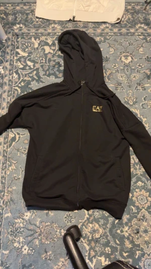 Svart EA7 hoodie med guldtryck - Svart hoodie från EA7 Emporio Armani med guldtryck på bröstet och en stor cirkulär guldlogga på ryggen. Tröjan har huva med snörning, hel dragkedja och två fickor framtill. Perfekt för dig som gillar streetwear och snygga detaljer.