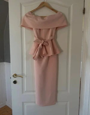 Ljusrosa långklänning med volang - Säljer en elegant ljusrosa långklänning med offshoulder och bred krage. Klänningen har ett snyggt midjeband som knyts till rosett och en volang upptill som ger extra stil. Perfekt för dig som vill sticka ut med en feminin och chic look.
