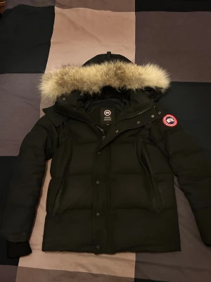 Svart Canada Goose dunjacka med päls - Säljer en svart dunjacka från Canada Goose med fluffig päls på huvan och ikonisk röd logga på ärmen. Jackan har dragkedja, knappar och flera fickor. Perfekt för kalla vinterdagar och har en riktigt schysst look.