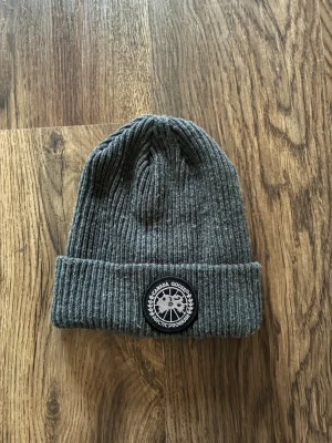 Grå ribbstickad mössa Canada Goose - Snygg grå ribbstickad mössa från Canada Goose med uppvikt kant och klassisk svartvit logotyp framtill. Perfekt accessoar för kalla dagar och enkel att matcha med olika outfits. Tillverkad i mjukt och värmande material. Pris kan diskuteras vid snabb affär