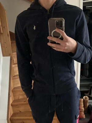 Mörkblå hoodie från Ralph Lauren - Säljer en mörkblå hoodie från Ralph Lauren med dragkedja och huva. Tröjan har klassisk vit logga på bröstet, känguruficka och ribbade muddar. Perfekt för chill dagar och enkel att matcha med det mesta. Den är small men passar en 185 man. 
