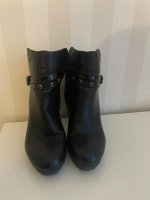 Svarta boots med klack från Renato Balestra - Snygga svarta boots från Rieker med hög klack och rund tå. Skorna har ett dekorativt bälte med nitar och spänne runt ankeln samt dragkedja på sidan. Tillverkade i skinnimitation med blank detalj. Perfekt för dig som gillar edgy stil.