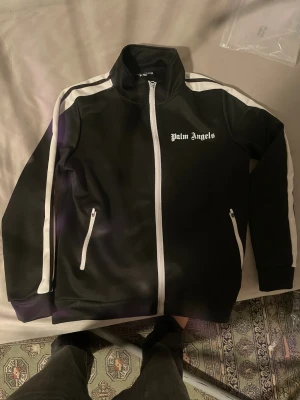 Palm Angels svart track jacket - Svart track jacket från Palm Angels med vita ränder längs ärmarna och vit dragkedja. Jackan har hög krage, två fickor med dragkedja och Palm Angels-logga på bröstet. Snygg och sportig design som sticker ut.