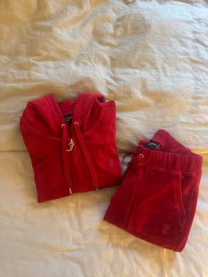 Juicy Couture set - Juicy Couture röd hoodie i mjuk velour med huva och snörning. Dragkedjan har en snygg J-detalj i silver och tröjan har broderad logga på bröstet. Perfekt för dig som gillar klassisk 00-talsstil och vill ha något riktigt comfy.