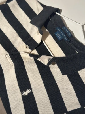 Randig långärmad pikétröja Ralph Lauren - Snygg långärmad pikétröja från Ralph Lauren med breda svarta och vita ränder. Klassisk krage, knappar framtill och broderad logga på bröstet. Tröjan är gjord i mjuk bomull och har en normal passform som funkar till många olika stilar.