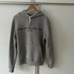 Grå hoodie Tommy Hilfiger - Snygg grå hoodie från Tommy Hilfiger med logotyp och broderad detalj framtill. Tröjan har huva med vit snörning