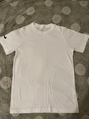 Vit Nike t-shirt med logga - Vit t-shirt från Nike i storlek L, med klassisk rund hals och korta ärmar. T-shirten har en svart Nike Swoosh-logga tryckt på vänster ärm. Tillverkad i mjuk bomull som känns skön mot huden. Enkel och stilren design som funkar till det mesta.