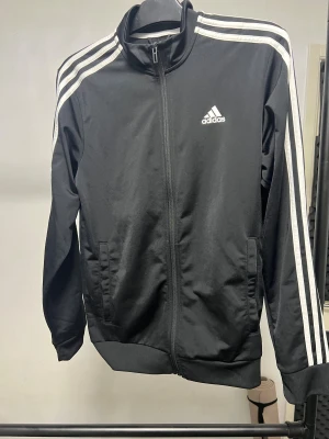 Svart Adidas track jacket med vita ränder - Svart Adidas track jacket med klassiska vita ränder längs ärmarna och vit logga på bröstet. Jackan har hög krage, hel dragkedja framtill och ribbade muddar. Tillverkad i polyester för en sportig vibe. Perfekt för dig som gillar streetwear och sportig stil.